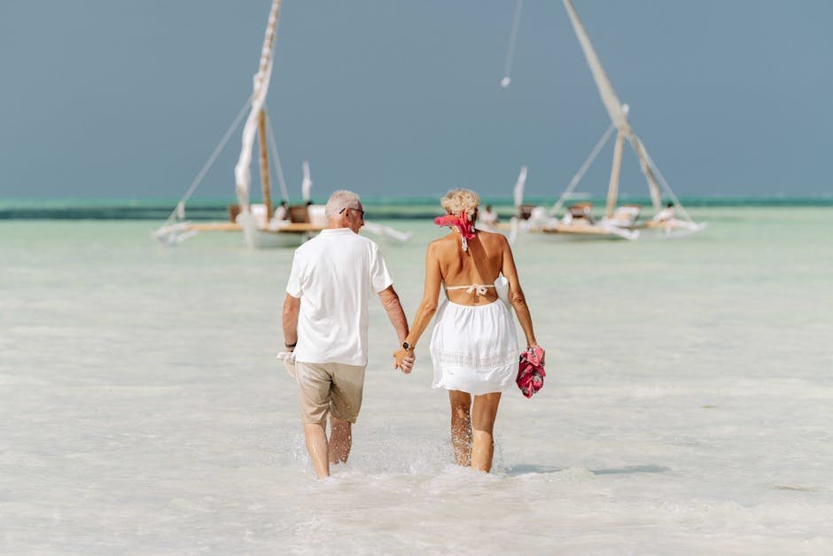 An elderly couple holds hands while walking on the beach, with dhows in the background in Zanzibar.
